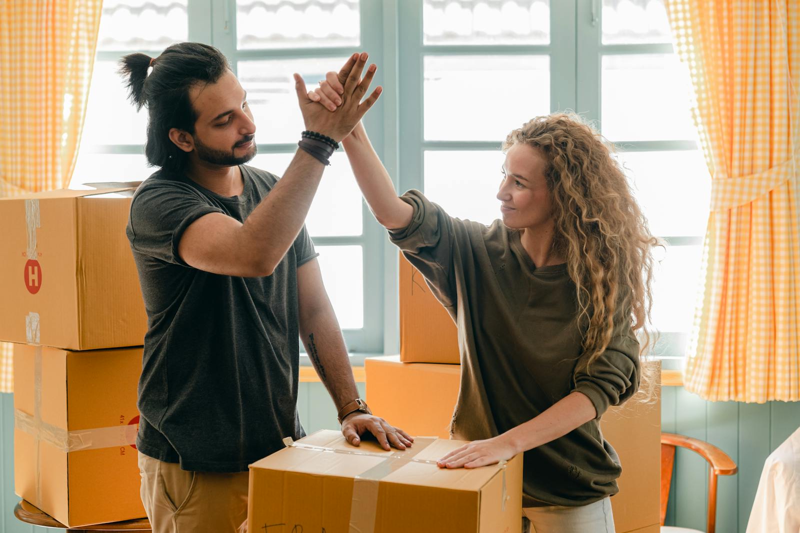 renters Happy woman in casual wear standing near heap of cardboard boxes and giving high five to ethnic boyfriend with ponytail showing agreement while looking at each other